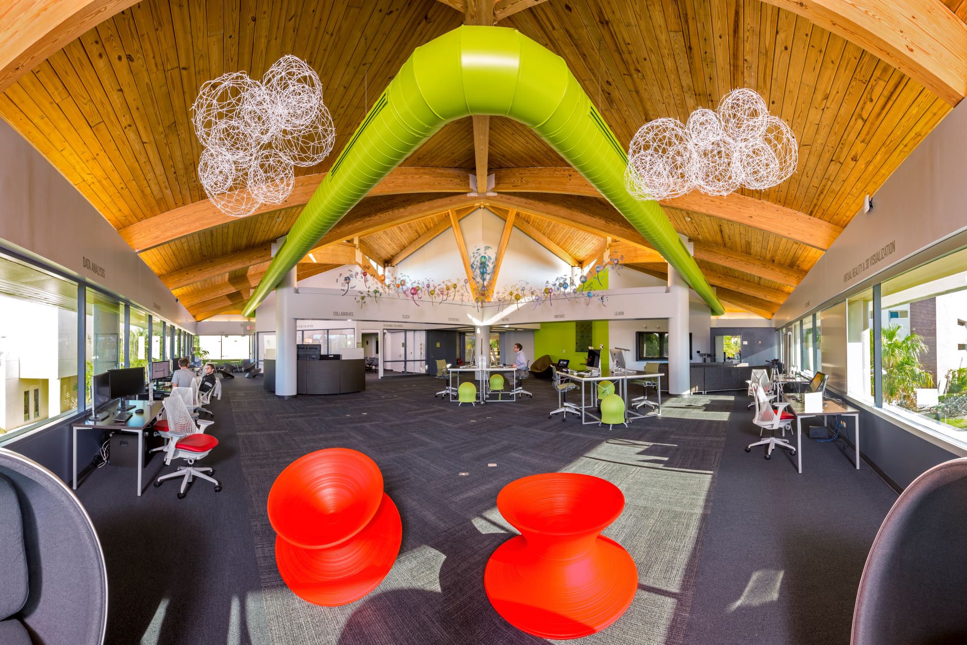 Modern, vibrant university study lounge with wooden ceilings, green structural beams, whimsical white light fixtures, and bright red lounge chairs. The space is equipped with various workstations and collaboration areas under natural lighting